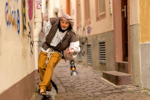 Ein verkleideter Schauspieler in einer dynamischen Pose als Freiburger Bösewicht Neunfingerle in einem historischen Gässle mit fließendem Bächle.