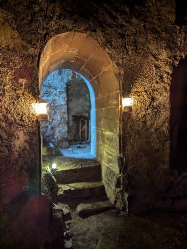 Ein düsterer, atmosphärischer mittelalterlicher Keller in Freiburg mit einer offenen Tür, durch die geheimnisvolles Licht in den historischen Raum fällt.